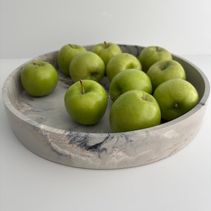 May include: A circular tray with a grey and white marble effect, holding ten green apples. The apples are a bright green and have stalks. The tray has a decorative design with touches of gold.