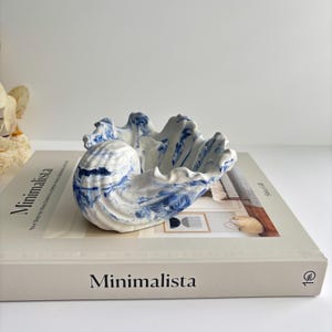 Könnte beinhalten: Eine keramische Muschelschale mit weißem und blauem Marmormuster. Die Schale liegt auf einem Buch mit dem Titel "Minimalista". Das Innere der Schale ist weiß mit blauen Streifen, und die Außenseite hat ein strukturiertes, wellenförmiges Design.