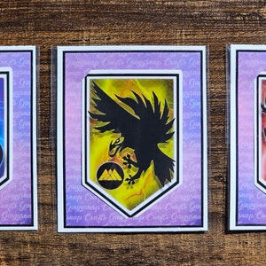 May include: Three framed art prints featuring stylized crest designs. One crest has a snake on a blue lightning background, another has a raven on a yellow background, and the last has a lion on a red background.