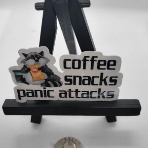 May include: A white sticker with a raccoon holding a bag of chips and the text "coffee snacks panic attacks". The sticker is on a small black easel.