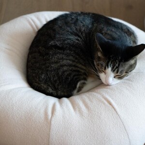 May include: A sleeping tabby cat, with black and grey stripes, curled on a light pink pet bed. The cat's face is white and grey, and its ears are perked. The bed is round and made of a soft, plush material.