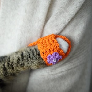 May include: A cat's paw is covered in a bright orange knitted mitten with a purple flower sewn on. The cat is resting on a soft, light beige sweater.
