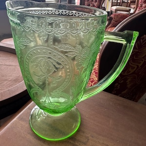 May include: A green glass pitcher with an embossed floral design. The pitcher has a handle and a wide base.