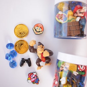 May include: Two clear plastic jars filled with various colorful plastic toys, including Mario, Luigi, Donkey Kong, and gold coins. There are also blue gems, a black video game controller, and a red and yellow Mario figurine on a white surface.