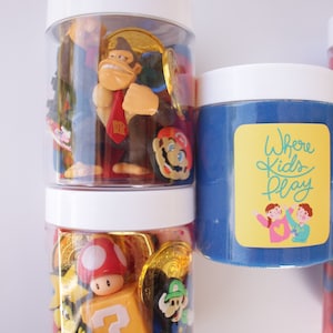 May include: Two clear jars filled with various colorful toys, including a red and white mushroom, a question mark block, and a Donkey Kong figurine. The jar on the right has blue slime and a yellow sticker that says "Where Kids Play".