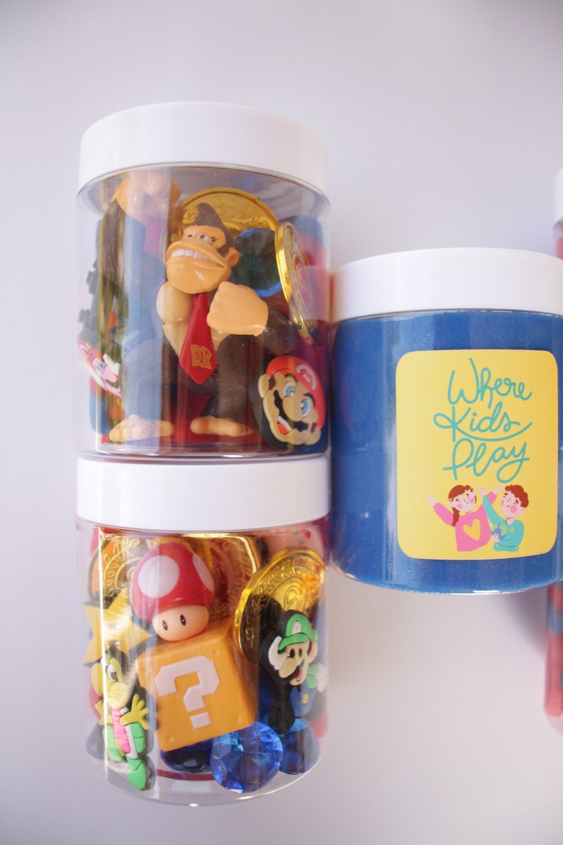 May include: Two clear jars filled with various colorful toys, including a red and white mushroom, a question mark block, and a Donkey Kong figurine. The jar on the right has blue slime and a yellow sticker that says "Where Kids Play".