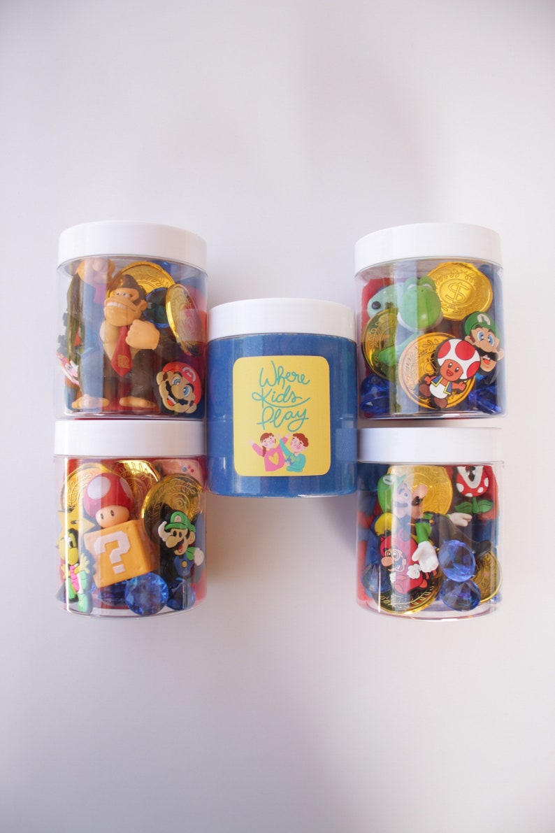 May include: Five clear jars filled with colorful toys and coins. The jars have white lids. The middle jar has blue slime and a label that says "Where Kids Play".