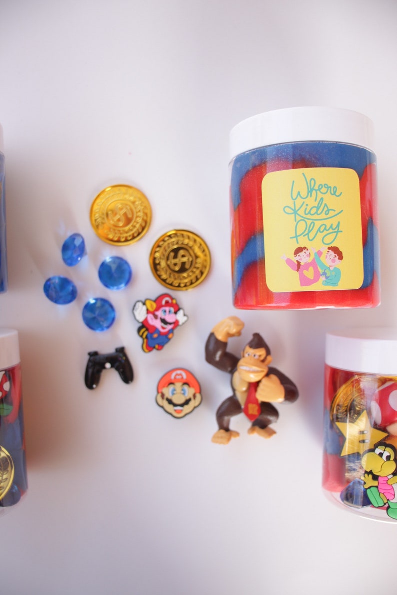 May include: Two containers of slime with a red and blue swirl pattern. The containers have white lids and labels that say "Where Kids Play". The slime is surrounded by various small toys, including gold coins, blue gems, a video game controller, and Mario characters.