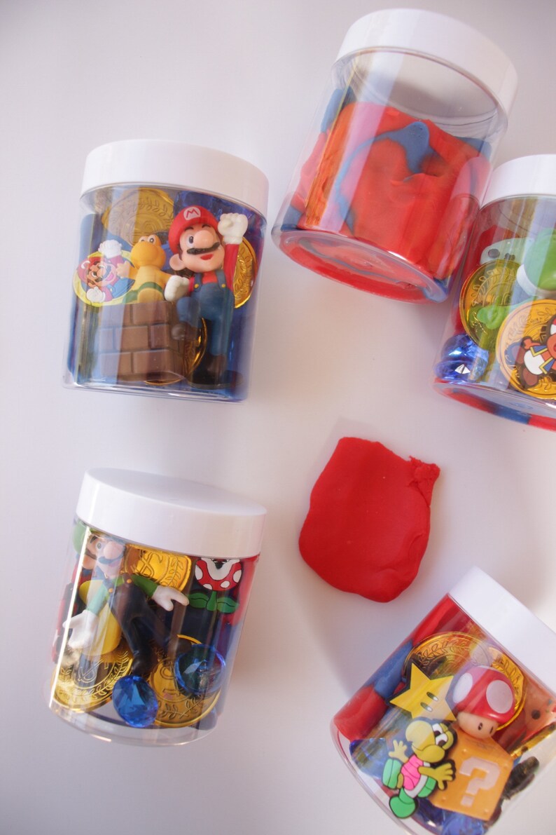 May include: Five clear plastic jars with white lids filled with various small toys and trinkets. The jars contain red and blue play dough, gold coins, blue gems, and small plastic figures of Mario and Luigi characters from the Super Mario video game series.