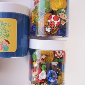 May include: Two clear plastic jars with white lids filled with blue gems, gold coins, and various Mario characters. The jars are on a white surface. The text "Where Kids Play" is on the label of the jar on the left.
