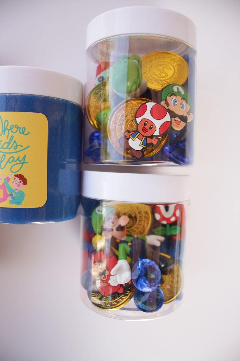 May include: Two clear plastic jars with white lids filled with blue gems, gold coins, and various Mario characters. The jars are on a white surface. The text "Where Kids Play" is on the label of the jar on the left.