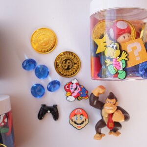 May include: A clear plastic jar filled with various colorful plastic toys, including a red mushroom, a yellow star, a blue question mark, and a brown monkey figurine. The jar is sitting on a white surface with blue and gold coins, a blue gem, a black video game controller, and a red and blue Mario figurine.