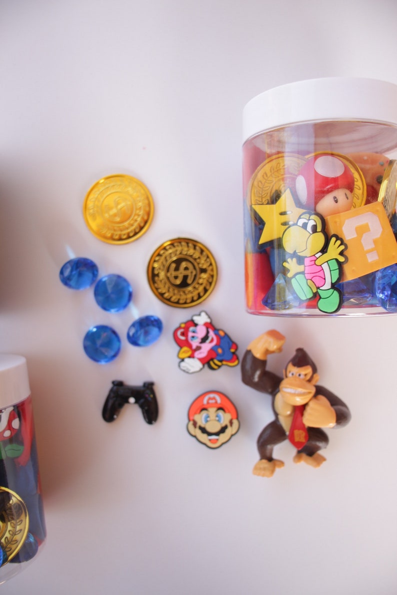 May include: A clear plastic jar filled with various colorful plastic toys, including a red mushroom, a yellow star, a blue question mark, and a brown monkey figurine. The jar is sitting on a white surface with blue and gold coins, a blue gem, a black video game controller, and a red and blue Mario figurine.