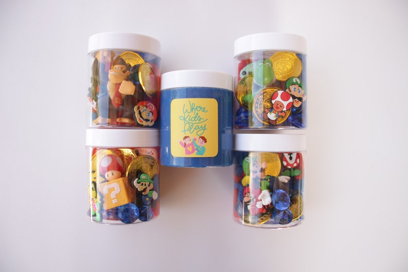 May include: Five clear jars with white lids filled with various colorful plastic toys, including Mario, Luigi, and other characters from the Super Mario Bros. video game franchise. The jars are arranged in a row on a white surface. The middle jar contains blue slime with a yellow label that reads "Where Kids Play".