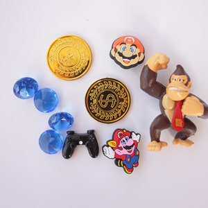 May include: A collection of video game themed toys and accessories, including a gold coin with a dollar sign, a blue gem, a black video game controller, a red and blue Mario character, a brown Donkey Kong character, and a red and white Mario character.