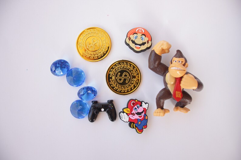May include: A collection of video game themed toys and accessories, including a gold coin with a dollar sign, a blue gem, a black video game controller, a red and blue Mario character, a brown Donkey Kong character, and a red and white Mario character.
