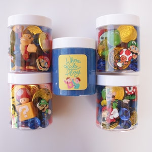 May include: Five clear jars with white lids filled with various colorful plastic toys, including Mario characters, coins, and gems. The middle jar contains blue slime with a yellow label that reads "Where Kids Play".