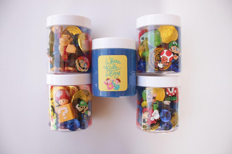 May include: Five clear jars with white lids filled with various colorful plastic toys, including Mario characters, coins, and gems. The middle jar contains blue slime with a yellow label that reads "Where Kids Play".