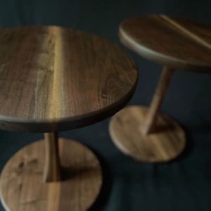 May include: Two wooden end tables with a dark brown finish. The tables have a round top and a single, slender leg.