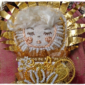 May include: A handmade embroidered angel figurine with a gold halo, white beads, and a pink and gold dress. The angel is surrounded by gold foil and white beads.