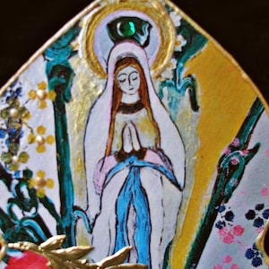 May include: A colorful, hand-painted, ceramic wall hanging featuring a depiction of the Virgin Mary. The artwork is in a teardrop shape and features a gold background with a green, blue, and white floral design.