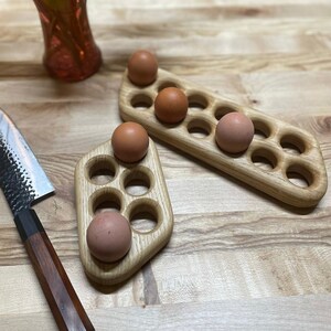 May include: Two wooden egg holders, one with six slots and one with five slots, with brown eggs in some of the slots. The holders are on a light wood surface.