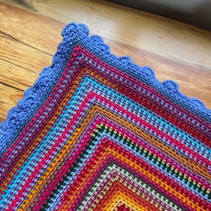 May include: A colourful crocheted blanket with a scalloped edge. The blanket is made with a variety of colours, including red, orange, yellow, green, blue, and purple.