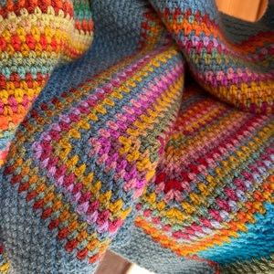 May include: A colorful crocheted blanket with a blue, green, yellow, orange, red, and pink color scheme. The blanket is made with a variety of stitches and textures.
