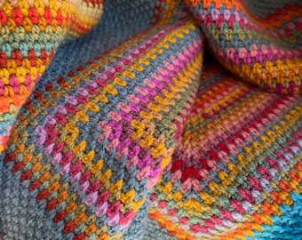 Può includere: Una coperta all'uncinetto colorata con uno schema di colori blu, verde, giallo, arancione, rosso e rosa. La coperta è realizzata con una varietà di punti e trame.