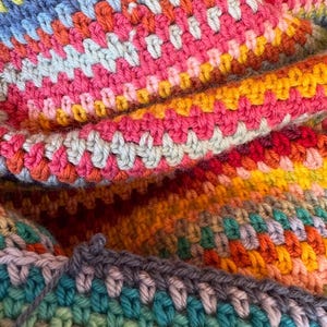 Puede incluir: Primer plano de una colorida manta de ganchillo hecha a mano. La manta presenta una vibrante mezcla de colores que incluyen rosa, naranja, amarillo, azul y verde. Las puntadas texturizadas crean un patrón visualmente atractivo.