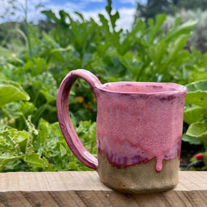 May include: A handmade ceramic mug featuring a textured, earthy brown base and a vibrant, glossy pink glaze that drips down the sides. The mug has a thick, curved handle and an irregular, organic rim shape, showcasing a distinct, hand-thrown appearance.