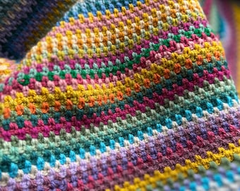Può includere: Una coperta all'uncinetto colorata con strisce di filato giallo, blu, verde, rosa e viola.