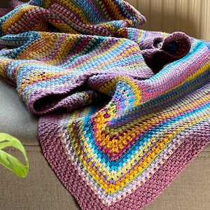 Puede incluir: Una manta de crochet colorida con rayas de color morado, amarillo, azul y verde. La manta está doblada y drapeada sobre un sofá marrón.