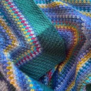 May include: A close-up of a crocheted blanket with a variety of colors, including blue, green, yellow, and pink. The blanket is made with a textured stitch and has a fringed edge.