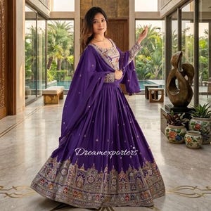 May include: A striking purple traditional dress with detailed embroidery and embellishments. The dress includes a long skirt, a matching shawl, and an ornate bodice. The text "Dreamexporters" is visible on the skirt. The dress is worn in a bright, luxurious setting.