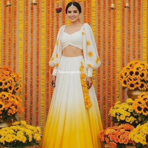 Op de afbeelding: Een vrouw draagt een witte en gele ombre rok en top met bloemaccenten. De top heeft lange mouwen met gele bloempatronen. De achtergrond is een muur van oranje en gele bloemen en slingers.