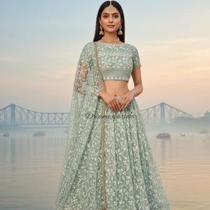 May include: A pale green lehenga with detailed white floral embroidery. The outfit comprises a matching short-sleeved top and a sheer dupatta. The lehenga has a wide, embroidered border. The text "Dreamexporters" is visible on the garment.