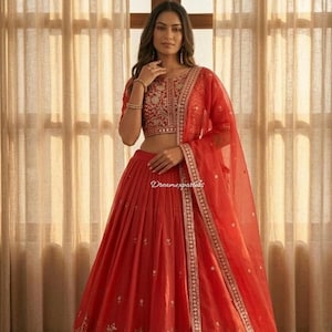 May include: A vibrant red lehenga, a traditional Indian garment, featuring intricate embroidery and embellishments. The ensemble includes a matching blouse and a sheer dupatta. The skirt has detailed floral patterns near the hem, and the blouse has detailed embroidery.