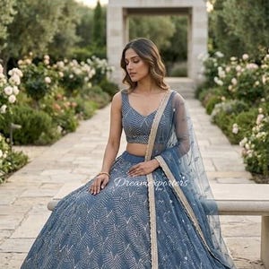 May include: A slate blue lehenga set featuring a sleeveless choli and a voluminous skirt, both adorned with intricate silver chevron embroidery. A matching sheer dupatta with a gold-toned border and delicate floral embellishments completes the set.