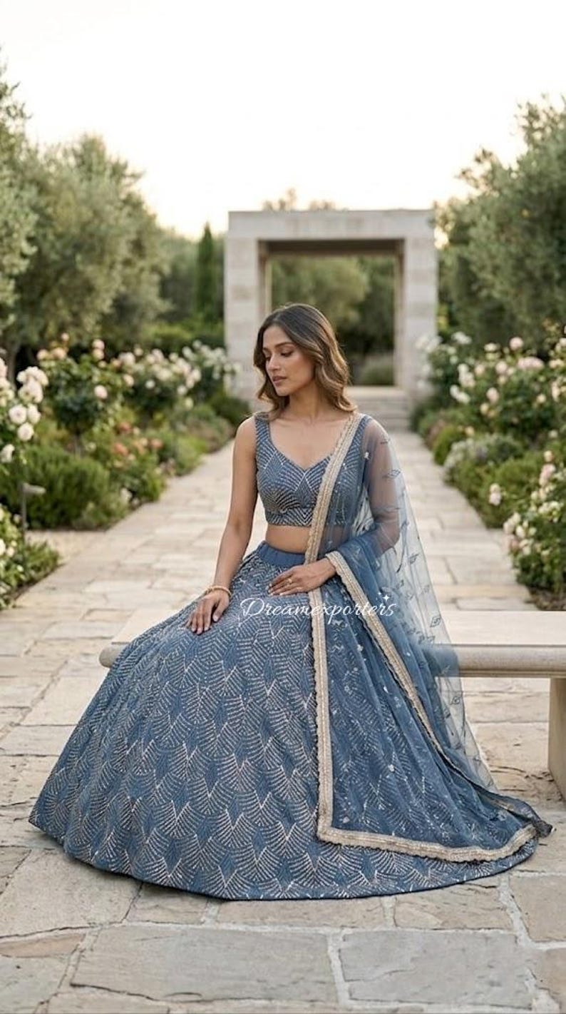May include: A slate blue lehenga set featuring a sleeveless choli and a voluminous skirt, both adorned with intricate silver chevron embroidery. A matching sheer dupatta with a gold-toned border and delicate floral embellishments completes the set.