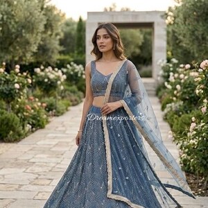 May include: A slate blue lehenga set featuring a sleeveless choli and a floor-length skirt, both adorned with intricate silver embroidery in a fan-like pattern. A matching sheer dupatta with a gold-toned border drapes over the shoulder.