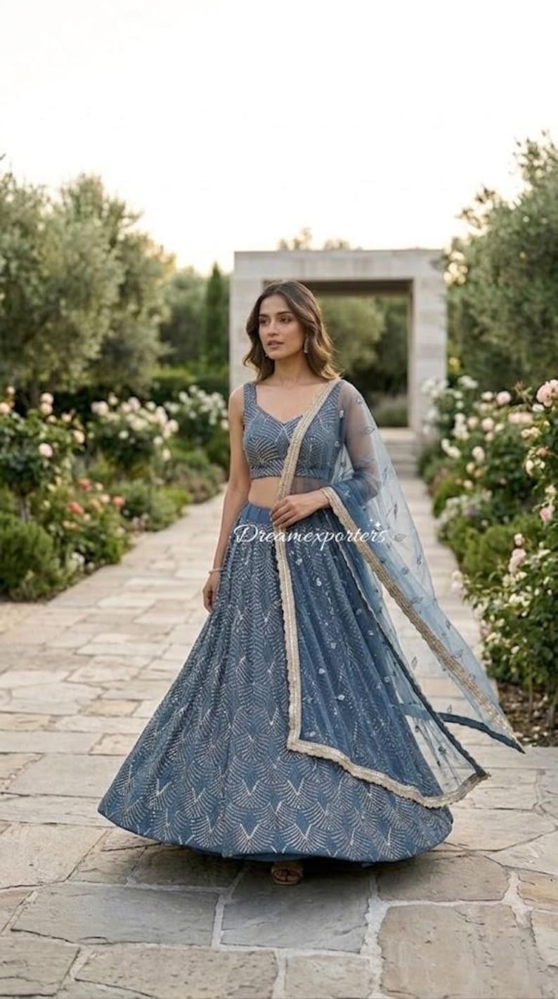 May include: A slate blue lehenga set featuring a sleeveless choli and a floor-length skirt, both adorned with intricate silver embroidery in a fan-like pattern. A matching sheer dupatta with a gold-toned border drapes over the shoulder.