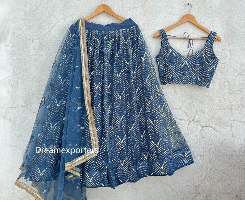 Puede incluir: Un conjunto lehenga azul con un top corto y una dupatta a juego. La falda y el top est&aacute;n adornados con un patr&oacute;n repetitivo dorado y blanco. La dupatta tiene un borde dorado. El conjunto est&aacute; colgado en perchas de madera.