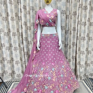 Puede incluir: Un lehenga bordado en rosa y blanco con un dupatta rosa a juego. El lehenga tiene un diseño floral con flores rosas, azules y verdes. El dupatta está drapeado sobre el hombro y tiene un diseño floral similar.
