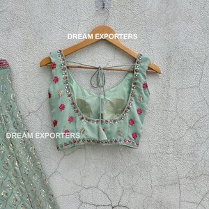 Op de afbeelding: Een lichtgroen, mouwloos blouse met een roze en gouden geborduurde ontwerp. De blouse heeft een strik aan de achterkant en hangt aan een houten hanger. De tekst "DREAM EXPORTERS" is zichtbaar op de hanger.