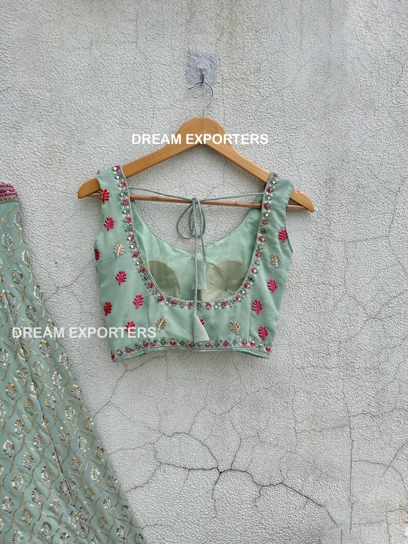 Op de afbeelding: Een lichtgroen, mouwloos blouse met een roze en gouden geborduurde ontwerp. De blouse heeft een strik aan de achterkant en hangt aan een houten hanger. De tekst "DREAM EXPORTERS" is zichtbaar op de hanger.