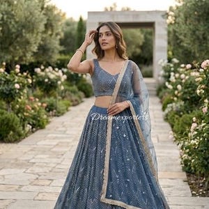 May include: A slate blue lehenga set featuring a sleeveless choli and a floor-length skirt, both adorned with a repeating silver chevron pattern. A matching sheer dupatta with a gold-toned border is draped over the shoulder.