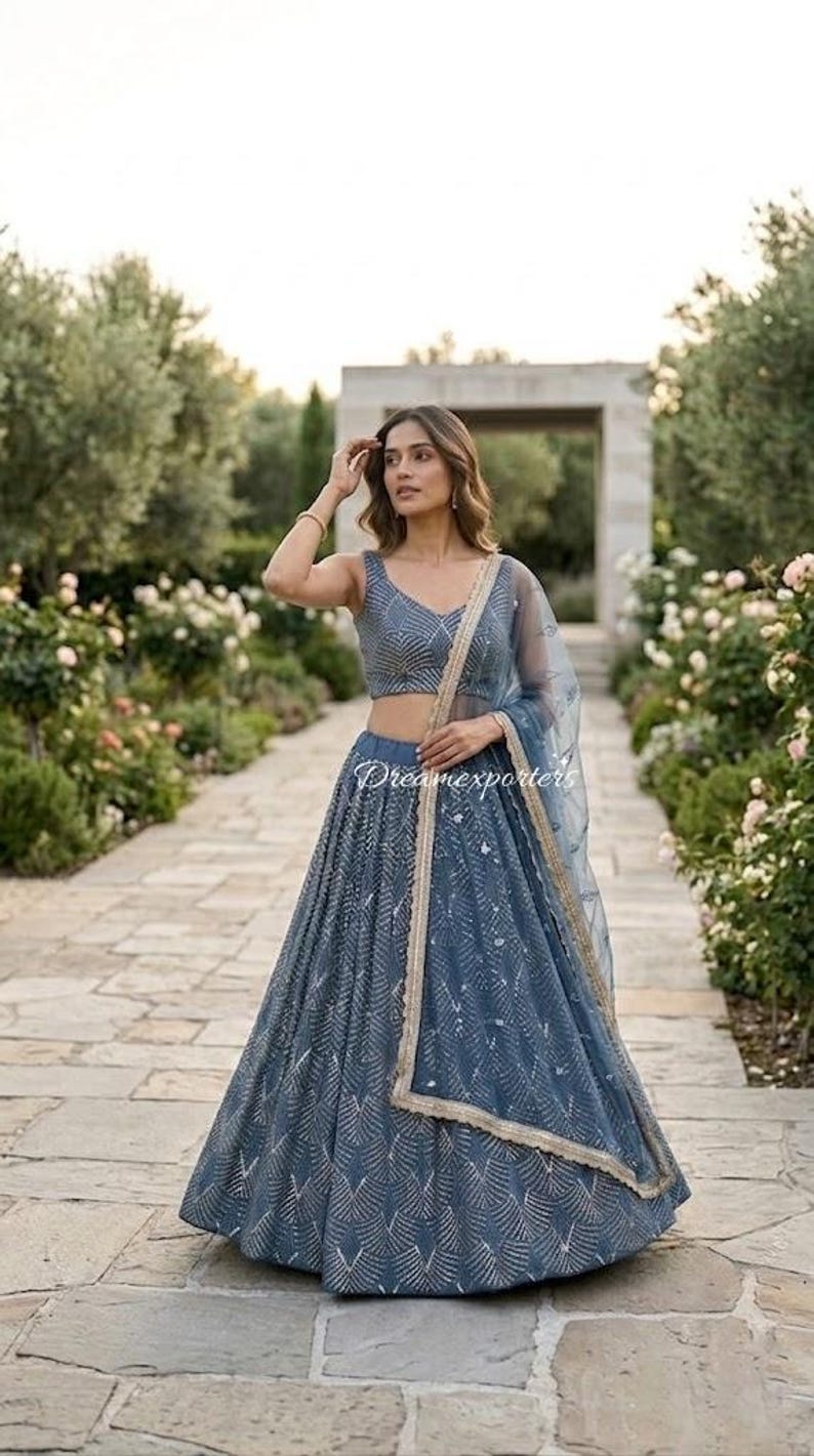 May include: A slate blue lehenga set featuring a sleeveless choli and a floor-length skirt, both adorned with a repeating silver chevron pattern. A matching sheer dupatta with a gold-toned border is draped over the shoulder.