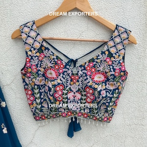 May include: A teal embroidered top with a floral design, featuring pink, red, and yellow accents. The top has a criss-cross back and is displayed on a wooden hanger. The text "DREAM EXPORTERS" is visible.