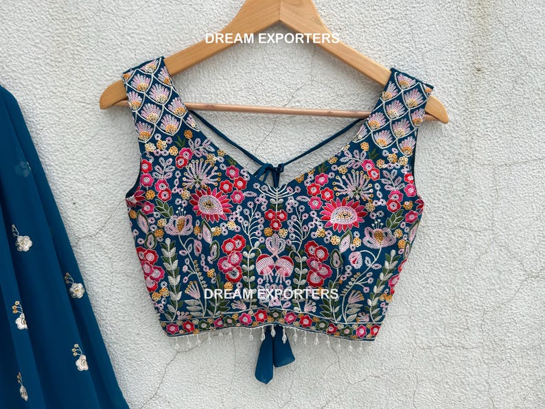 May include: A teal embroidered top with a floral design, featuring pink, red, and yellow accents. The top has a criss-cross back and is displayed on a wooden hanger. The text "DREAM EXPORTERS" is visible.